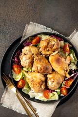 Fried chicken legs with salad in a plate on a dark kitchen table. Cooked poultry meat, pickles, and tomatoes on a culinary background top view	