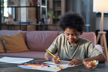 Naklejka premium Happy schoolboy of African ethnicity drawing picture with crayons while sitting by table in home environment