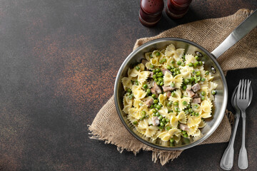 Farfalle with green peas, turkey ham and parmesan in cream sauce in a frying pan on the kitchen table. Traditional Italian pasta on a dark culinary background top view from the copyspace	