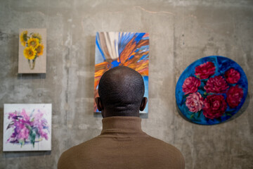 Rear view of young African man looking at collection of abstract paintings on wall while visiting...