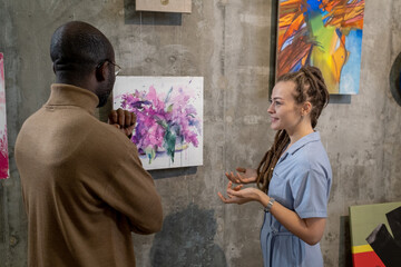 Young female guide describing one of paintings on walls to African man visiting modern art gallery