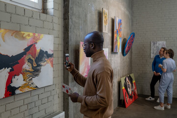 Young African male visitor of art gallery taking photo of painting on wall while enjoying...