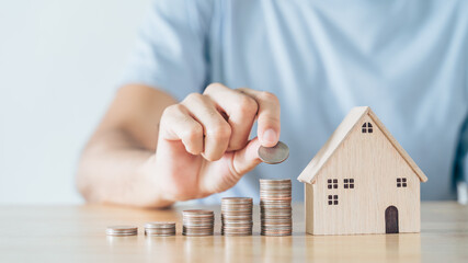 Man hand put coin on coins stack with wooden house on wood table. saving money for buying house,...