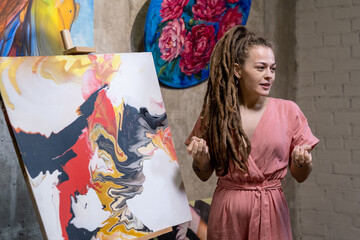 Young female auctioneer or speaker with dreadlocks describing abstraction illustrated on painting in front of audience