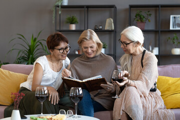 Mature friends sitting on sofa drinking wine and watching photo album together in the living room