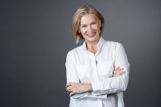Portrait of mature woman with short hair in white blouse standing with arms crossed and smiling at camera isolated on grey background