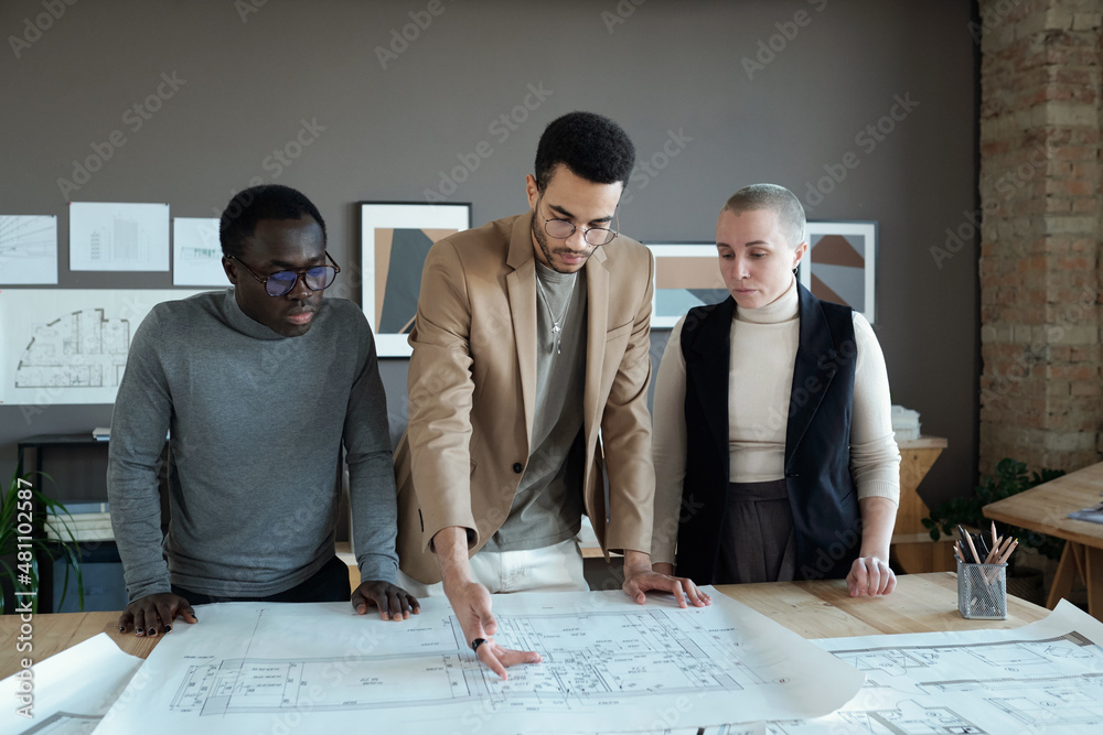 Poster young biracial businessman making presentation of sketch on unrolled blueprint to his colleagues at 