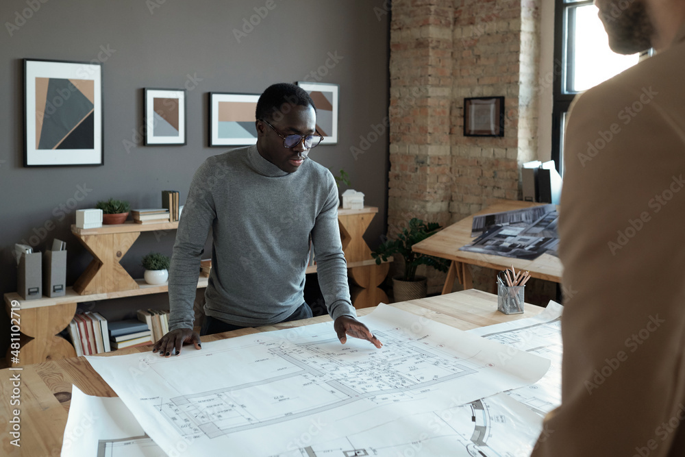 Poster young confident african engineer looking at sketch while discussing it with male colleague at workin