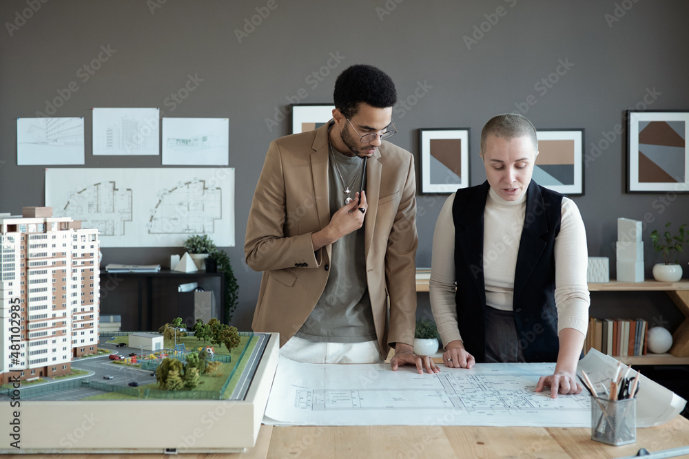 Poster Two elegant architects or engineers discussing sketch on blueprint while standing by large table with house layout