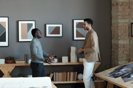 Two young intercultiral businessmen discussing working points while standing in front of each other against wall with paintings