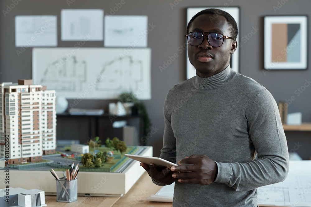 Poster professional engineer of african ethnicity scrolling in tablet while standing in office and looking 