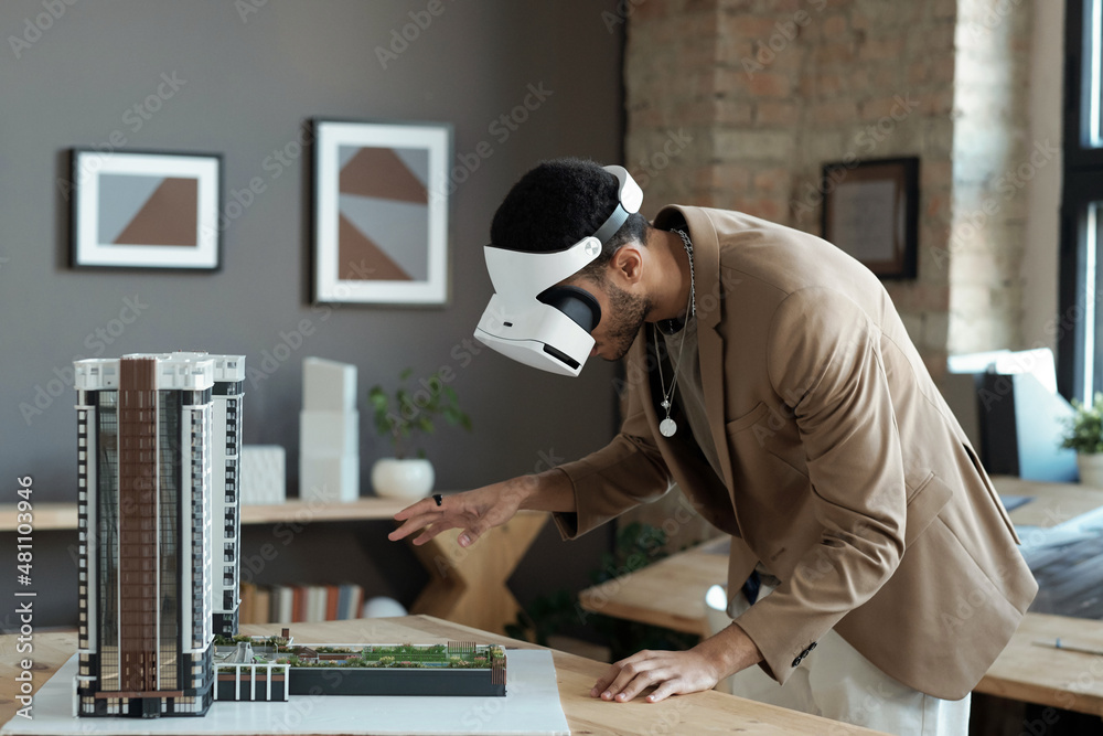 Poster Young architect in vr headset bending over house layout while standing by table and making presentation of new architectural project