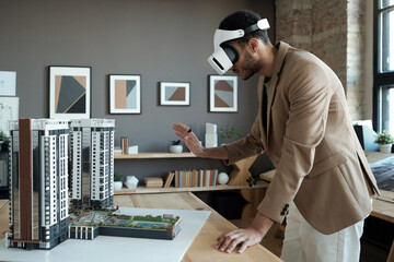 Young elegant architect in vr headset standing by table with house layout during presentation of...