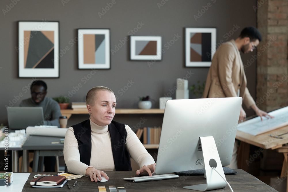 Poster serious mature female designer looking at computer screen while sitting by table and looking through