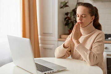 Indoor shot of relaxed woman with ponytail wearing casual style beige sweater sitting at table in...