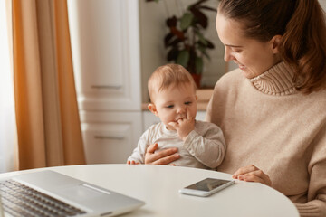 Indoor shot of beautiful Caucasian woman wearing beige casual style sweater with ponytail hairstyle sitting at table with her baby daughter and looking at baby with charming smile and gentle.