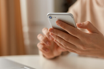 Indoor shot of unknown faceless woman wearing beige casual style sweater using mobile phone for checking social networks or typing messages, holing smart phone in hands.