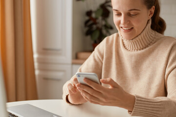 Indoor shot of smiling beautiful young woman wearing beige casual style sweater with ponytail hairstyle sitting at table and holding mobile phone in hands, checking social networks or writing post.