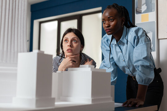 Project engineers women colleagues looking inspired in front of 3d residential maquette in architectural modern office. Focused professional architects team inspecting skyscraper white foam model.