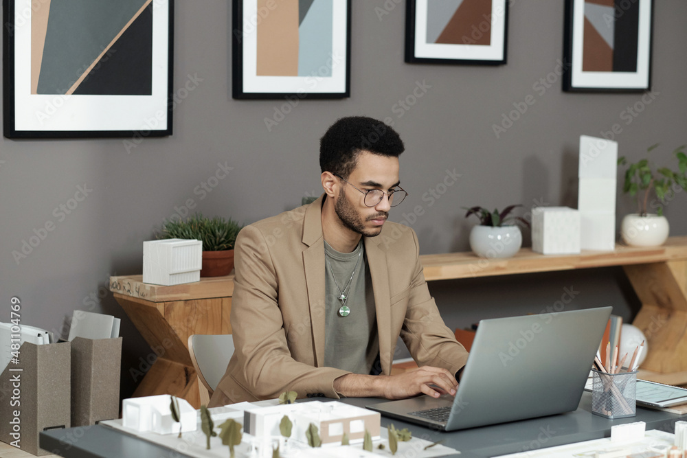 Poster young serious businessman in formalwear typing on laptop keypad while sitting by workplace and looki