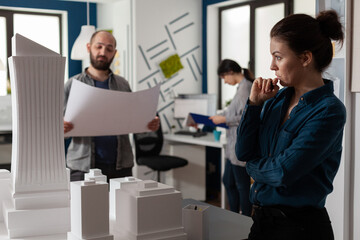 Architect looking into the distance gazing at skyscraper scale model with team colleague engineer holding construction plans. Team of architects brainstorming architectural ideeas.