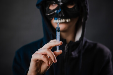 Close-up, selective focus on a syringe in the hands of a person wearing a skull mask and a hoodie,...