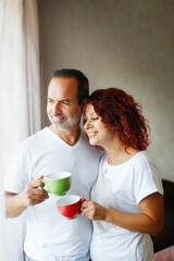 Cute middle aged european couple tender hugs and morning coffee together. Man and woman together at the window in the bedroom, lifestyle in a real interior. Support and emotions