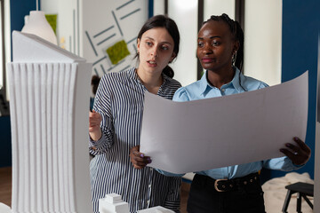 Architects couple looking at skyscraper 3d white foam scale model holding blueprints in modern architectural office. Team of two engineers discussing building construction plans.