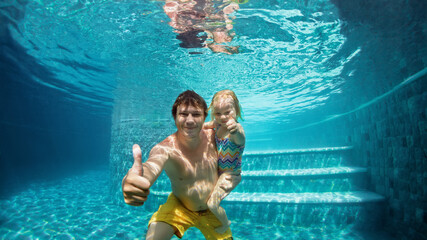 Happy people dive underwater with fun in aqua park swimming pool. Father, daughter show thumbs up....