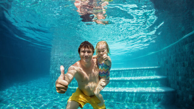 Happy people dive underwater with fun in aqua park swimming pool. Father, daughter show thumbs up. Family lifestyle, kids water sports activity, children swimming lesson with parents on summer holiday
