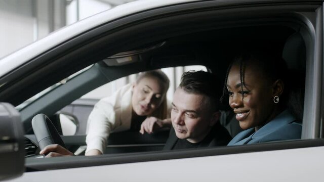Portrait of a smiling african lady and her european man inspecting the interior of a new car in a modern dealership. An attractive salesperson talks about an electric car and enjoys the beautiful car