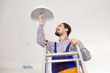 Electrician screwing in light bulb at home. Happy man in work uniform standing on ladder in residential building, office, school or hospital and changing LED lightbulb in modern white lamp on ceiling - obrazy, fototapety, plakaty