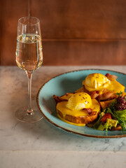 Breakfast in restaurant  - eggs Benedict with hollandaise cheese sauce served with glass of champagne wine in restaurant or cafe interior background. Close up, selective focus - 481114750