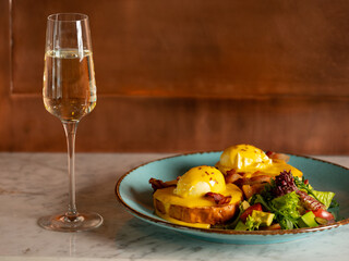 Breakfast in restaurant  - eggs Benedict with hollandaise cheese sauce served with glass of champagne wine in restaurant or cafe interior background. Close up, selective focus