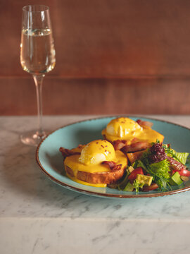 Breakfast In Restaurant  - Eggs Benedict With Hollandaise Cheese Sauce Served With Glass Of Champagne Wine In Restaurant Or Cafe Interior Background. Close Up, Selective Focus