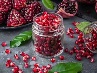 Glass jar with pomegranate seeds on a gray background surrounded by pomegranates and lemon leaves. Horizontal