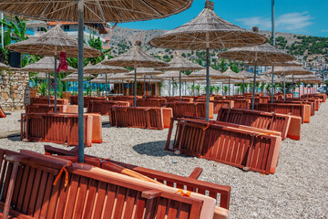 View of empty beach – wooden sunbeds and shade umbrellas near sea water, city buildings and mountains on the horizon. Landscape of abandoned summer resort.
