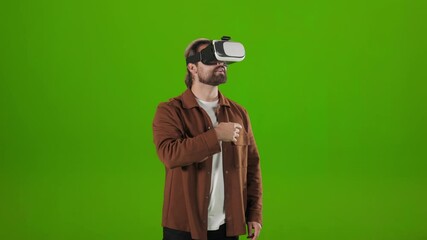 Caucasian man in VR headset standing over green background and gesturing with fingers in air. Bearded guy touching virtual screen in studio.