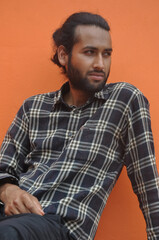 Fototapeta premium A good looking young man with beard and hair bun looking sideways while sitting outside, posing against orange wall background