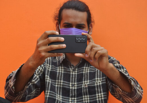 Selective focus of a young man wearing face mask with taking picture of you with his mobile phone, posing against orange color wall with copy space