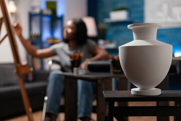 Selective focus on white vase standing on table for artist inspiration in art atelier. Young illustrator drawing sketch on canvas working at creative masterpiece using professional painting tools