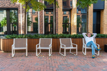 Millennial man in trousers and white T-shirt with long sleeves looking at camera while sitting on one of four chairs in courtyard of modern brick residential complex with landscaping on city street