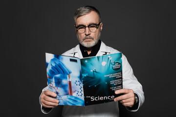 senior doctor in eyeglasses reading science magazine isolated on dark grey.