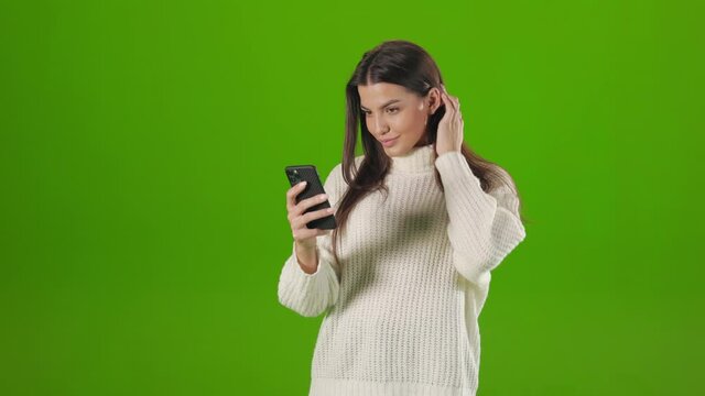 Charming young woman adjusting her long dark hair while taking selfie on modern smartphone. Positive brunette standing over green background with cell phone in hands.