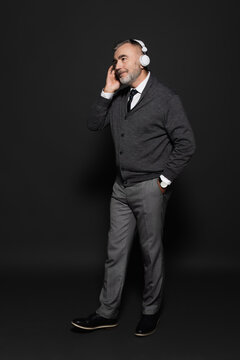 full length view of senior man in stylish clothes listening music in headphones on dark grey.