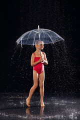 Beautiful little girl in red sportswear under umbrella in aqua studio. Drops of water spread about her fitness body. On the background of water splashes. Classic dark color background