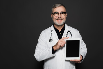 joyful senior doctor in white coat and eyeglasses holding digital tablet with blank screen isolated on black.