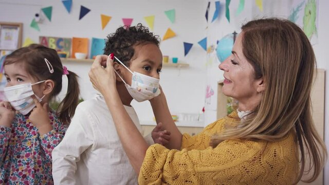 Pre School Teacher Helping Little Boy To Wear Face Mask Indoors At Nursery School, Coronavirus Concept.