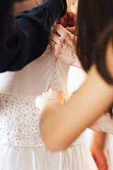 Obraz na płótnie Canvas Three bride assistants helping bride in white dress to prepare her dress back view. Morning bridal preparation details newlyweds. Wedding day moments, wear white dress. 