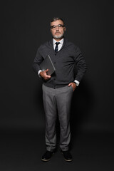 full length view of senior stylish man standing with hand in pocket and laptop on dark grey.